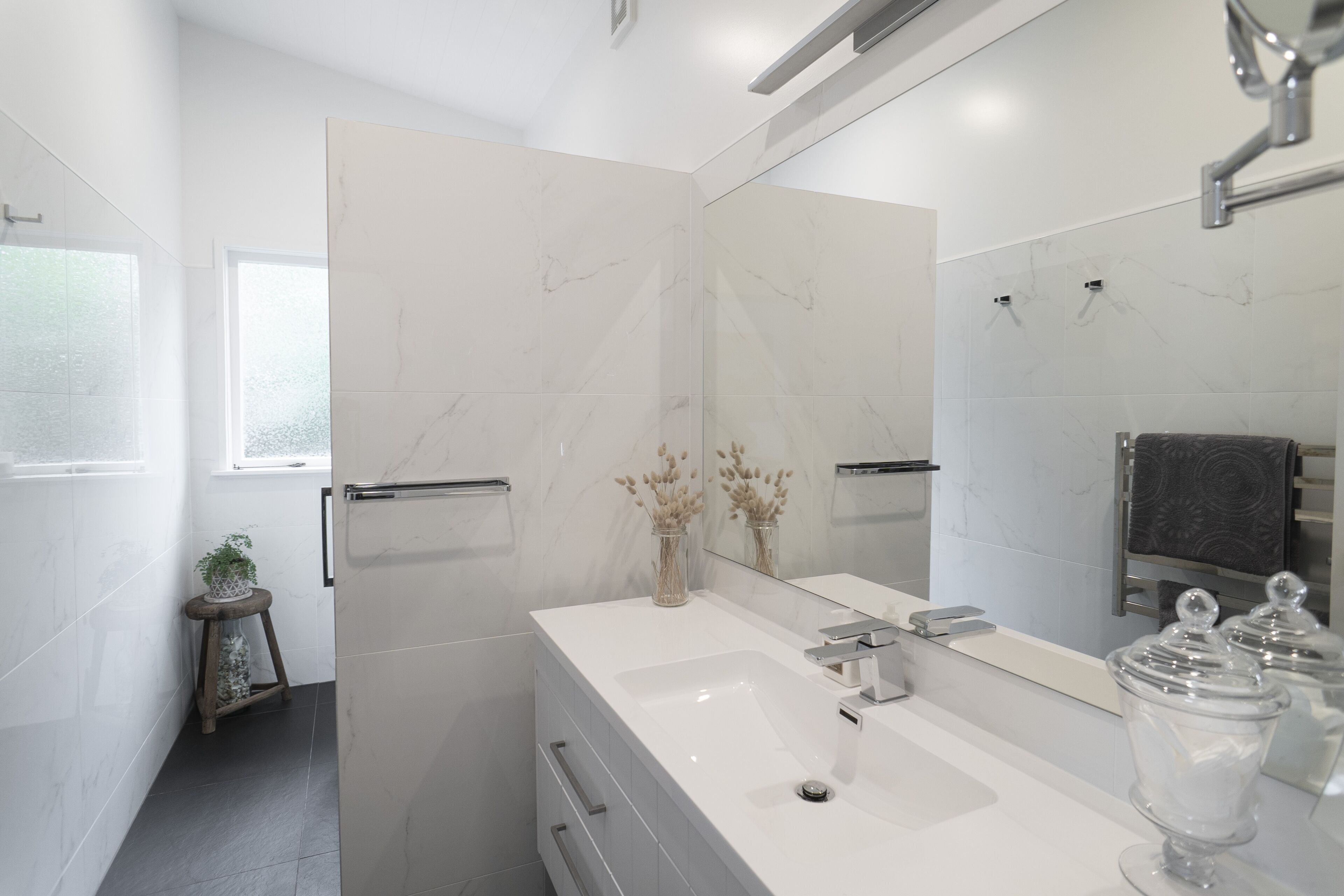 A modern bathroom with a large mirror, white marble tiles, a white sink, and a small plant on a wooden stool.