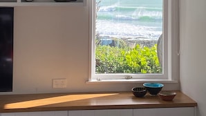 A kitchen with a window overlooking the ocean, a bookshelf, and a countertop with bowls. - Whale Bay Surf House + Spa - Absolute Waterfront (Waikato)
