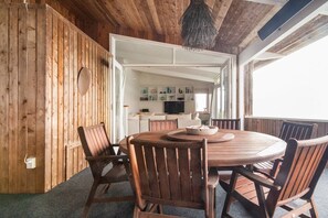 Et træinteriør med et rundt spisebord og stole, en hængende lampe, og udsigt til en opholdsstue med et fjernsyn og reoler.