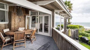 A wooden deck with a dining table and chairs, a hanging light fixture, and a view of the ocean. - Whale Bay Surf House + Spa - Absolute Waterfront (Waikato)