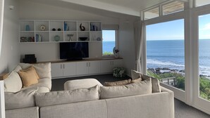 A living room with a large flat-screen TV, a white console table, and a sofa. There is a large window with a view of the ocean. - Whale Bay Surf House + Spa - Absolute Waterfront (Waikato)