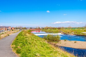 Een promenade langs de rivier met een sluisinstallatie, een pad langs een gazon, en gebouwen op de achtergrond.