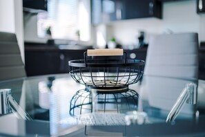 Een keuken met een glazen tafel, een metalen mand met een houten handvat en moderne apparaten op de achtergrond.