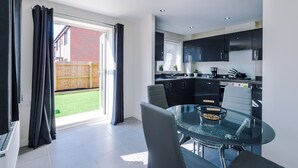 A modern kitchen with dark cabinets, a glass dining table, and a view of a backyard through a sliding door. - 3 Bedroom House with River Irwell view, sleeps 6 (Salford)