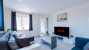 A modern living room with a flat-screen TV, a sofa, a coffee table, and a window with curtains. - 3 Bedroom House with River Irwell view, sleeps 6 (Salford)