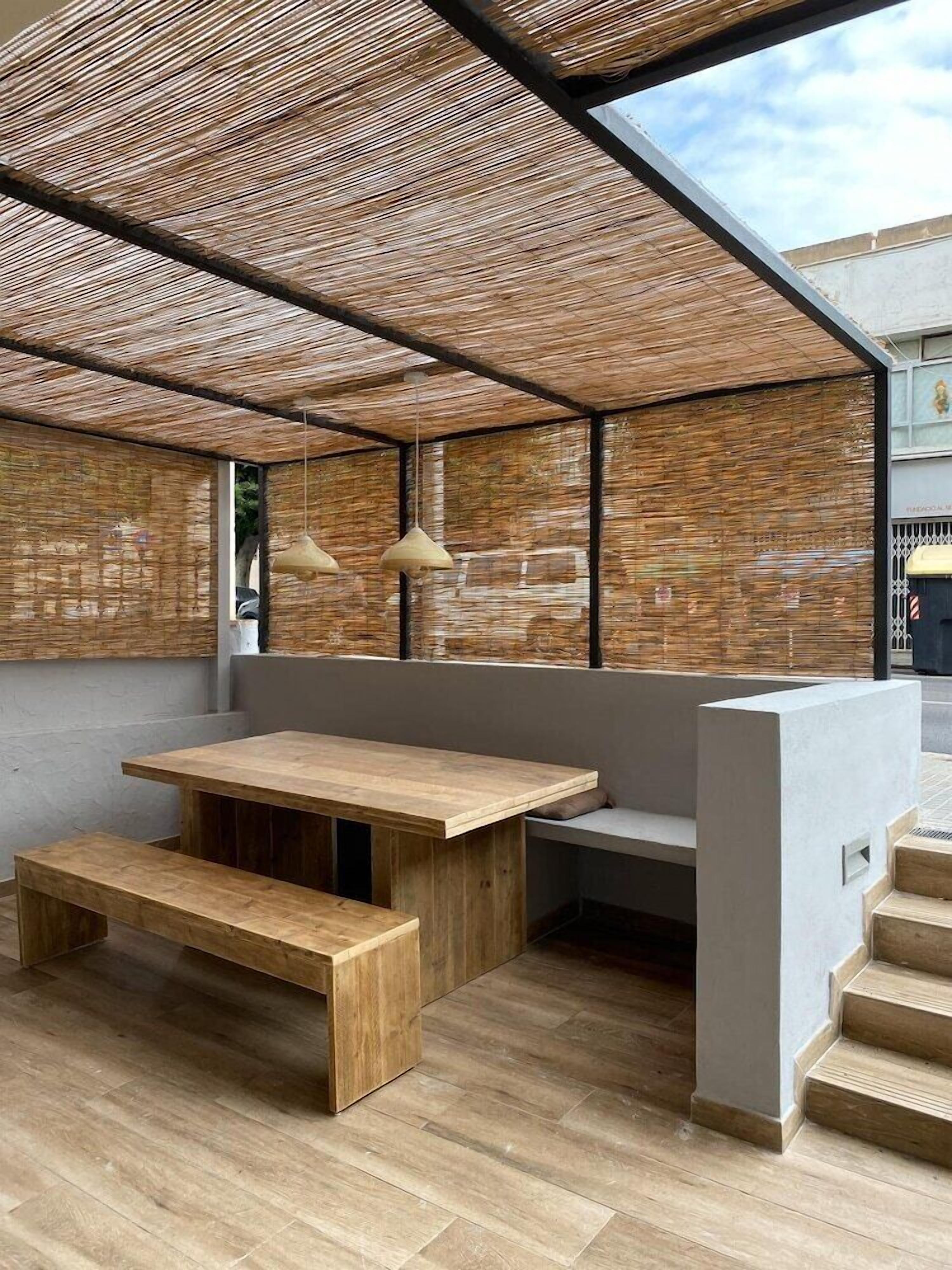 Een modern buiten zitgedeelte met een houten tafel en banken, een geweven zonnescherm en hangende lampen.