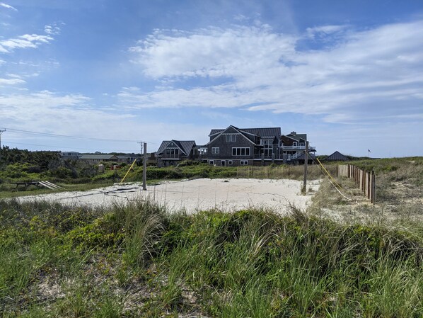 A large, dark-colored beachfront house with multiple levels and a prominent balcony, surrounded by a sandy beach and dense vegetation.