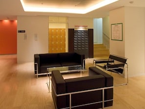 A modern lobby with a black leather sofa, a glass-top coffee table, and a metal-framed chair. There are lockers in the background and a staircase to the right. - Rhino (Kyoto)