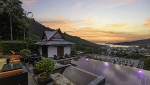 An infinity pool with a view of a city and hills, outdoor seating, and a small building with a sloped roof.