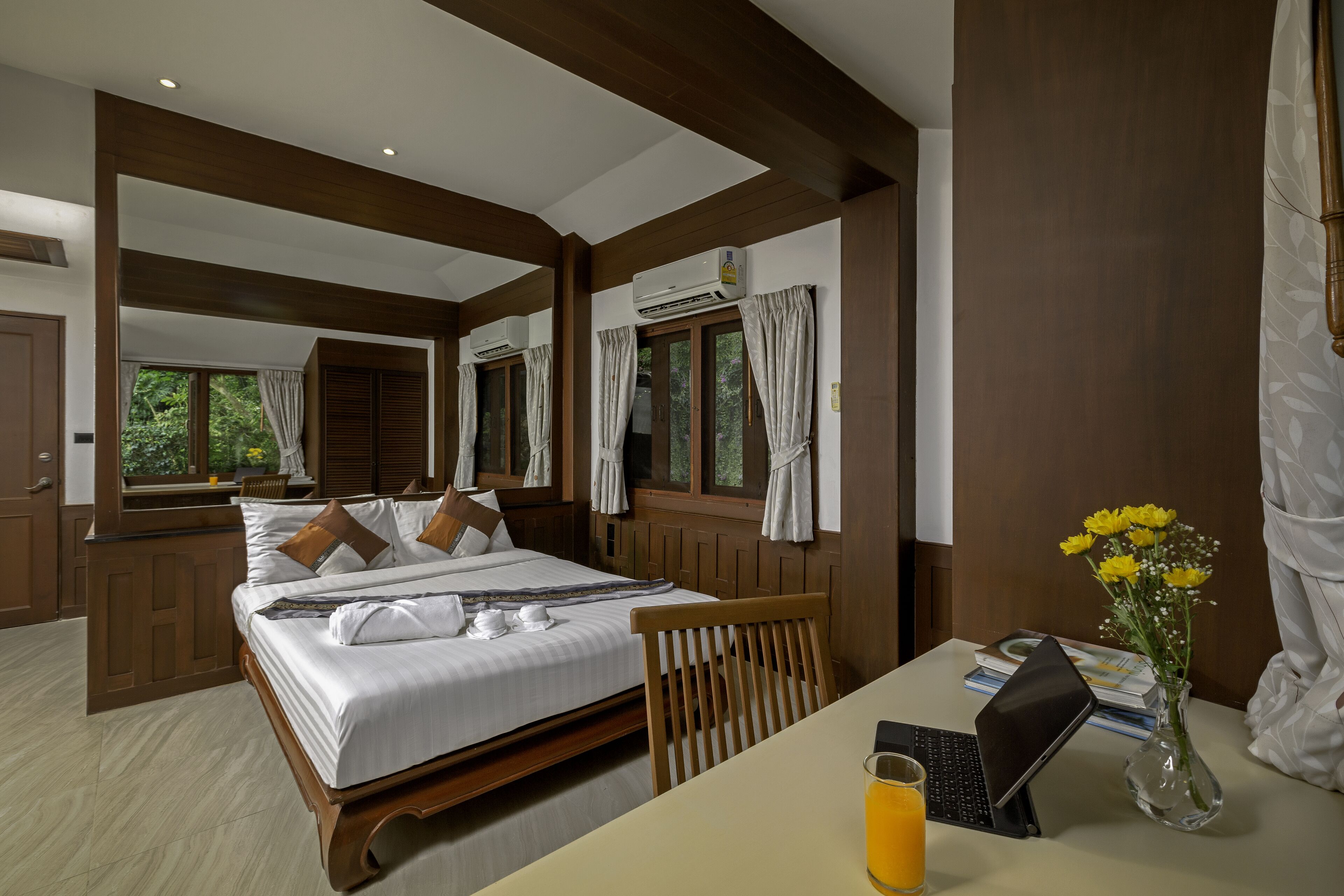 A hotel room with a large bed, a desk with a laptop, a vase with flowers, and a window with curtains.