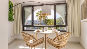 A room with a round table, two wicker chairs, a hanging light fixture, and a large window with curtains.