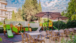 An outdoor patio area with wicker chairs and tables, a children's play structure, and a large umbrella providing shade. - Villa Aurora (Varna)