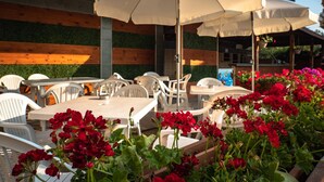 An outdoor seating area with white tables and chairs, surrounded by red geraniums and beige umbrellas. - Villa Aurora (Varna)