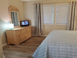Une chambre avec une commode en bois, un téléviseur posé dessus, des rideaux à motifs et un lit recouvert d’une couvre-lit à motifs géométriques.