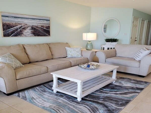Un salon avec deux canapés beige, une table basse blanche, un tableau représentant une plage, une lampe et un miroir.