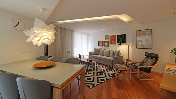 A modern living room with a dining area, featuring a wooden table, grey chairs, a patterned rug, a sofa, and a black leather chair. - FLH Sá da Bandeira Modern Apartment (Oporto)