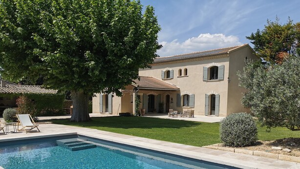 A swimming pool with a tiled deck, a large tree, and a building with a tiled roof and arched windows.