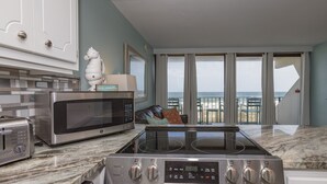 A modern kitchen with a stainless steel stove, microwave, and toaster. It features a large window with a view of the beach and a balcony with a railing. - Island Winds E205-Park The Car & Forget It! Location X3! OMG View! (Gulf Shores)