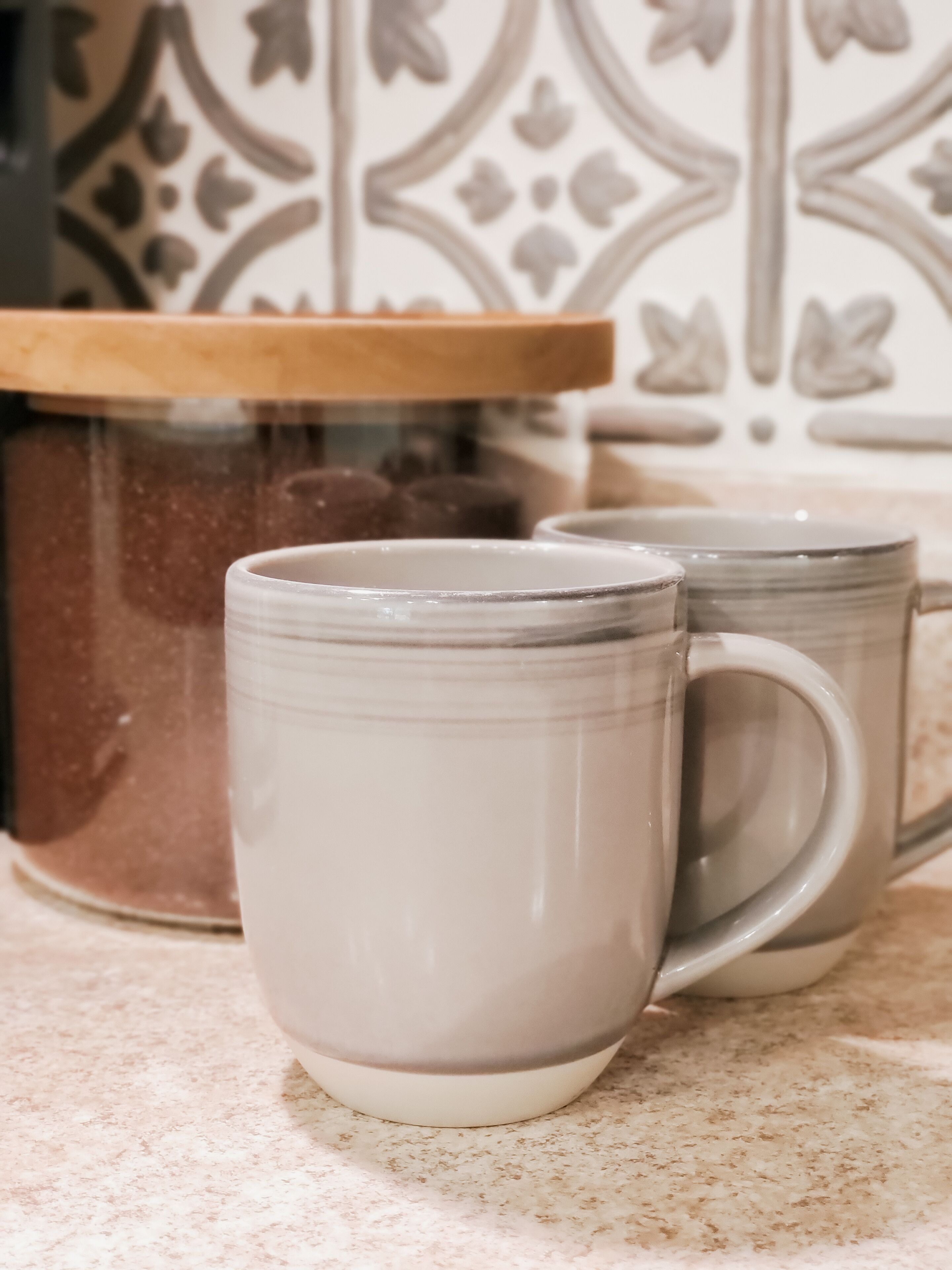 Deux tasses en céramique posées sur un comptoir, avec un mur recouvert de carreaux décoratifs.