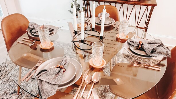 Una mesa redonda de vidrio dispuesta con platos, cubiertos y velas, rodeada de sillas de cuero marrón.