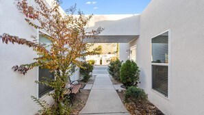 Un sendero estrecho que lleva a un edificio de fachada blanca, un pequeño árbol y una banca.