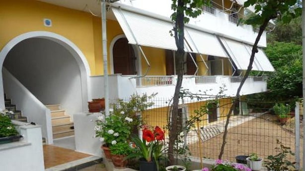 A two-story building with a yellow facade, white exterior walls, and a covered staircase. There are potted plants and a metal fence in the courtyard. - Anastasia Apartments (Corfu)