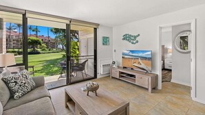 Un salon moderne doté d’une grande porte-fenêtre coulissante donnant sur un balcon, d’un téléviseur à écran plat, d’une table basse en bois et d’un canapé agrémenté de coussins à motifs.