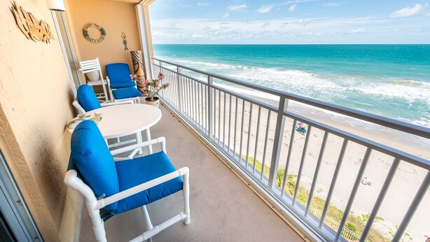 Varanda com cadeiras e mesa brancas, com vista para uma praia e ondas.
