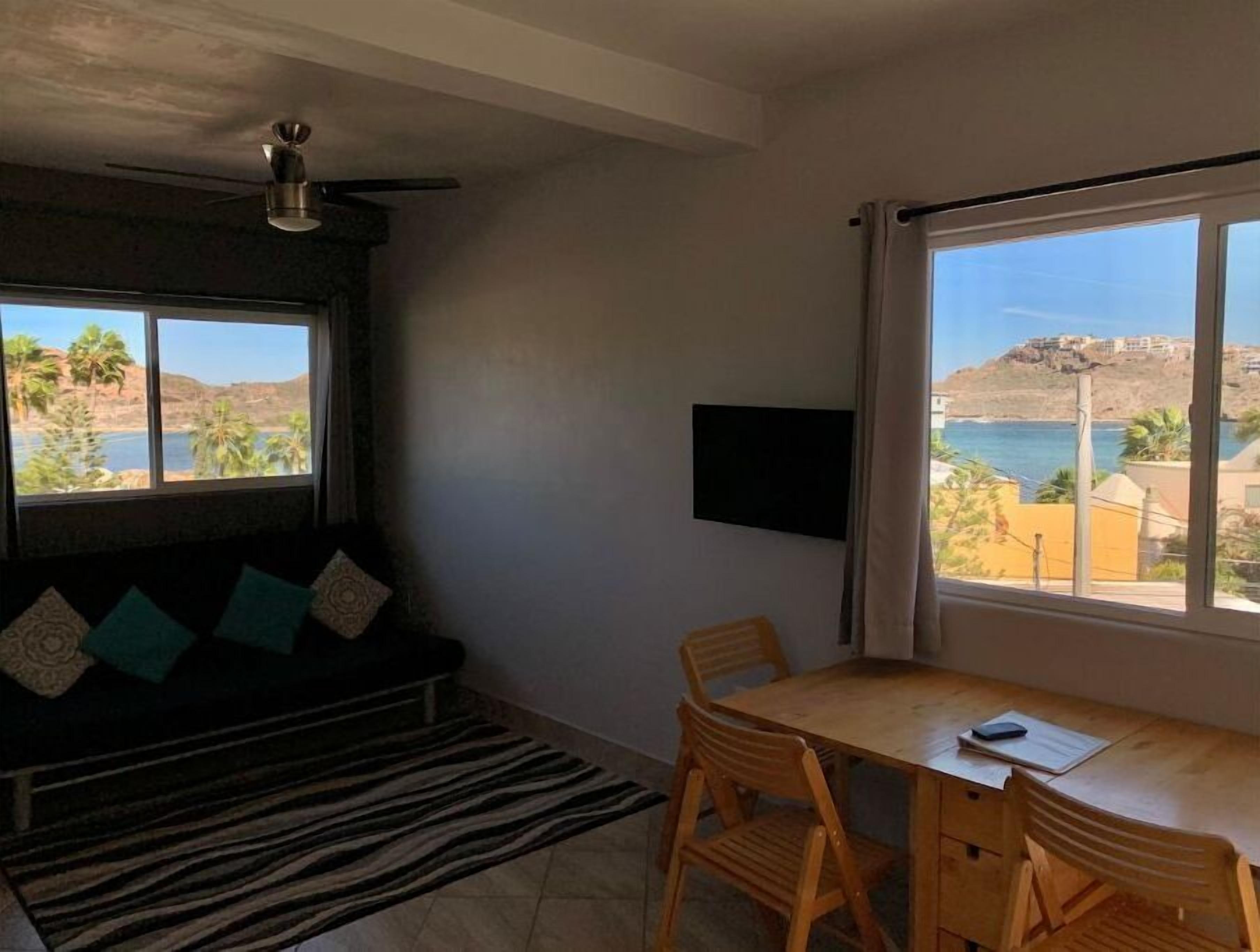 Una stanza con un divano, un tavolo da pranzo con sedie, una TV fissata al muro e ampie finestre con vista sul mare e sulle colline.