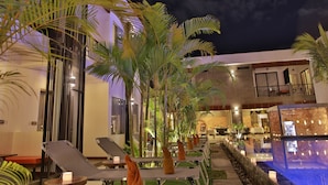 Zona junto a la piscina con sillones de descanso y palmeras, zona de bar con mesas y sillas, y un edificio con balcón.