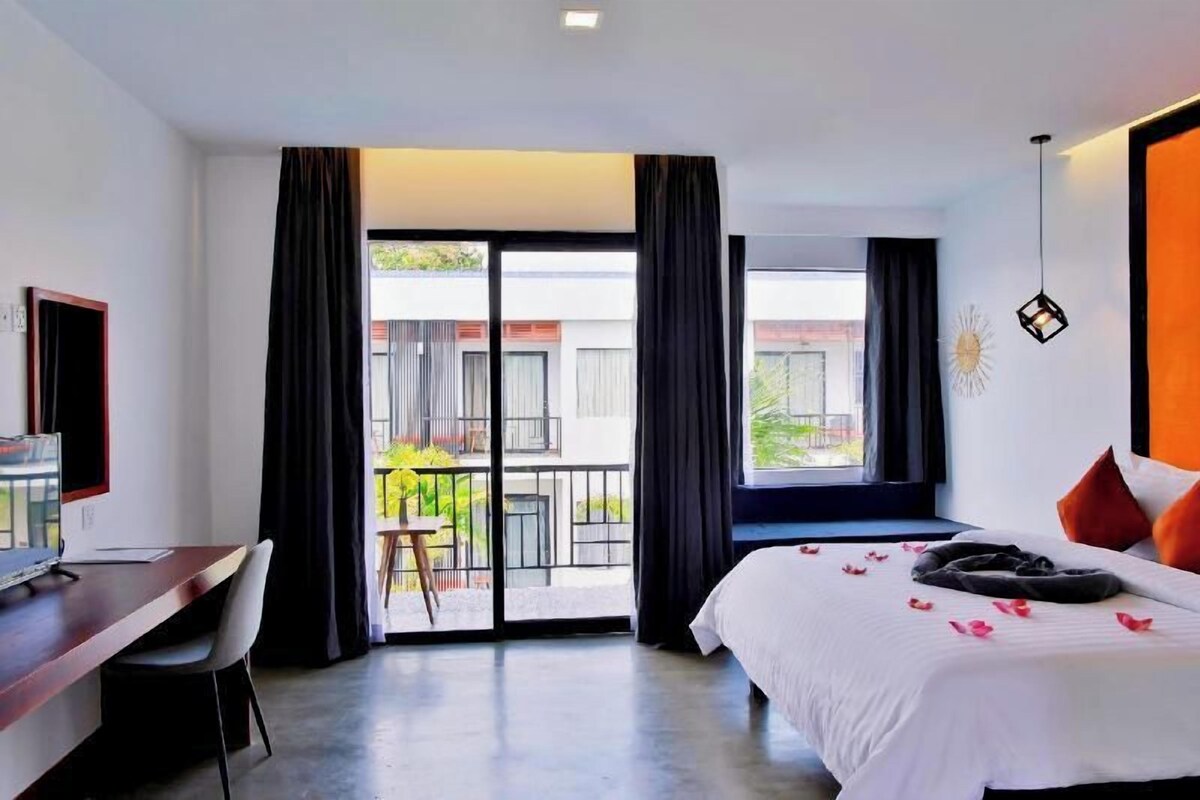 A hotel room with a bed, a desk, a chair, a TV, and a balcony with a view of buildings.