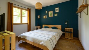 A bedroom with a wooden bed, a large window with curtains, a wall-mounted shelf, and framed pictures.