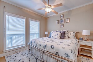 Une chambre à coucher comprenant un lit, deux fenêtres, un ventilateur de plafond et des œuvres d’art accrochées au mur.