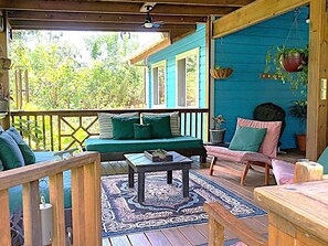 A covered porch with a blue exterior, wooden furniture, and a patterned rug.