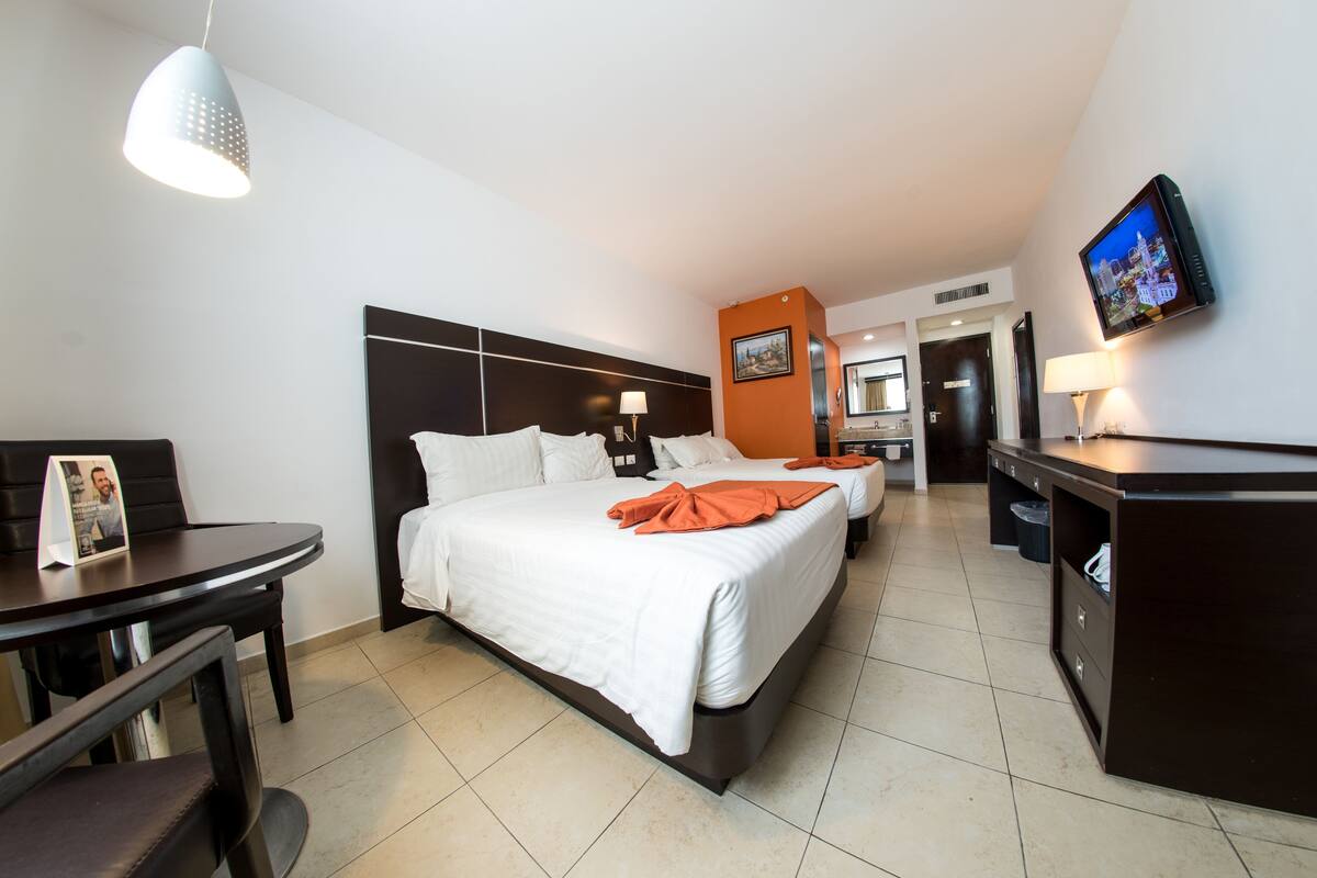 A hotel room with a large bed, a desk, a chair, a TV, and a bathroom visible in the background.