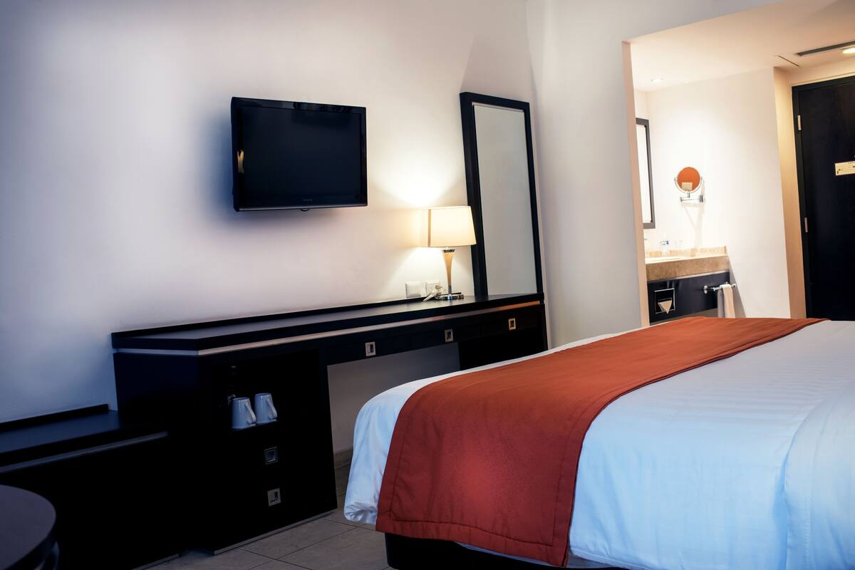 A hotel room with a bed, a desk with a flat-screen TV, a mirror, a lamp, and a bathroom visible through an open door.