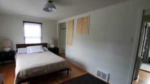 Un dormitorio con una cama, dos mesitas de noche, una ventana con persianas, una lámpara de techo y un radiador.