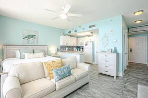 A bedroom with a bed, a white sofa, a ceiling fan, and a kitchenette in the background.