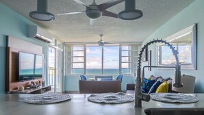A modern living room with a large TV, ceiling fan, and a view of the ocean through large windows. - Oceanfront Condo • Pool • Near Seven Seas Beach (Fajardo)