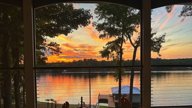 Uitzicht vanuit een kamer met grote ramen op een meer bij zonsondergang, met een steiger en een overdekt gedeelte.