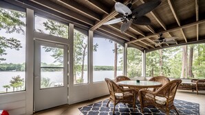 Een overdekt terras met een tafel en stoelen, grote ramen en uitzicht op een meer.