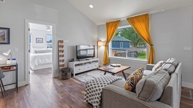 A modern living room with a sofa, TV, and a view of a blue house through large windows. - Pet & Family Friendly, Mountain Views, Downtown Montrose 20 Min to National Park (Montrose)