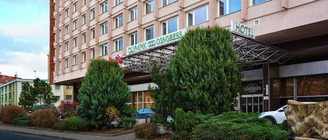 Un hotel con fachada moderna, un letrero que indica que es el Hotel del Congreso Olímpico, y un área de entrada cubierta con árboles y arbustos.