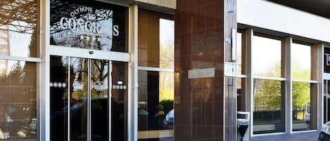 Une entrée d'immeuble avec des portes vitrées, une enseigne indiquant « Olympiapik Congress », et une zone pavée de briques.