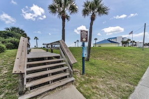 Una escalera de madera que conduce a una zona de playa con palmeras y un edificio al fondo.