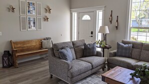 A living room with two grey sofas, a wooden bench, a small table with a lamp, and a large window with a view of greenery.