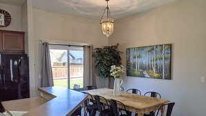 A dining area with a wooden table, black chairs, a chandelier, a painting of a forest, and a view through a sliding door to a backyard.
