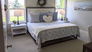 A bedroom with a bed, two bedside tables, a nightstand, a wall-mounted lamp, a decorative wreath, and a painting.