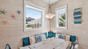Une pièce avec un mur en bois, une table à manger en bois avec des chaises, un banc avec des coussins, des décorations en forme d’étoiles de mer et un panneau indiquant les règles à respecter sur la plage.