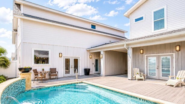 Une piscine avec une terrasse en bois, un patio couvert et un bâtiment à deux étages dont la façade est blanche.
